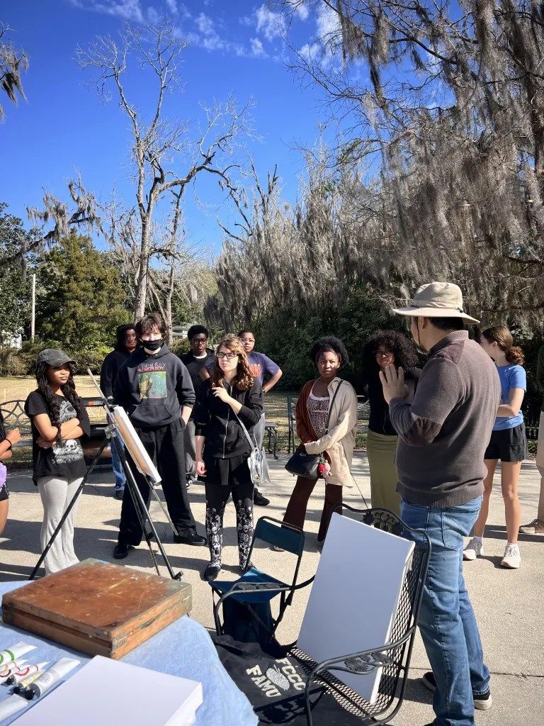 Outdoor group listening to a man in a cowboy hat beside easels and a table on a sunny path