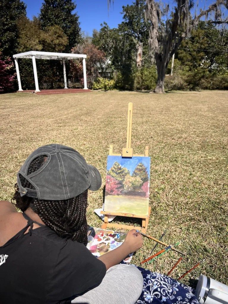 Person painting outdoors at an easel in a grassy park, with a white pavilion in the background.