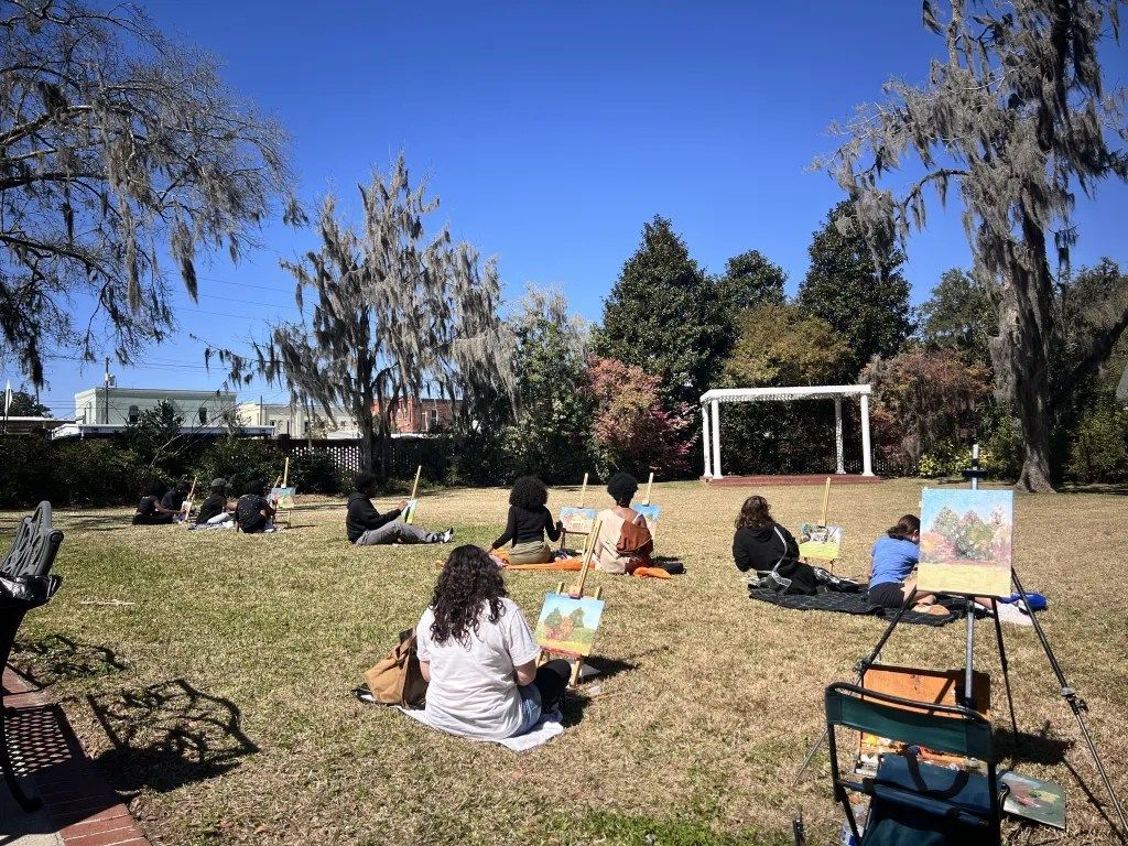 People painting outdoors on a grassy lawn under a blue sky, with easels and trees in the background.