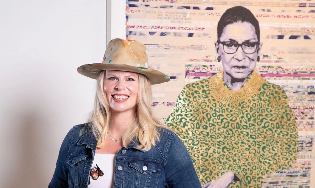Smiling woman in denim jacket and hat posing beside a life-size portrait cutout against a patterned wall
