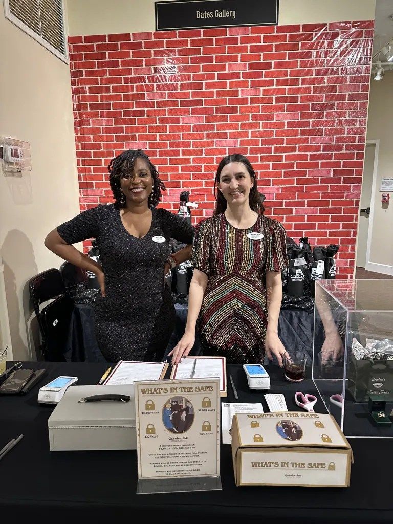 Two women behind a display table at a booth, with a red brick backdrop and event flyers in front.