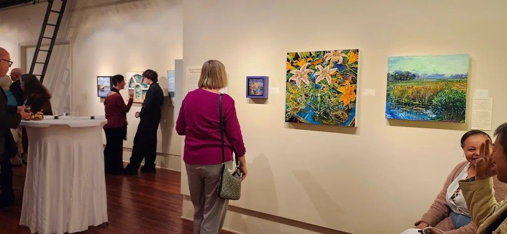 Art gallery with visitors viewing colorful landscape paintings on a white wall.
