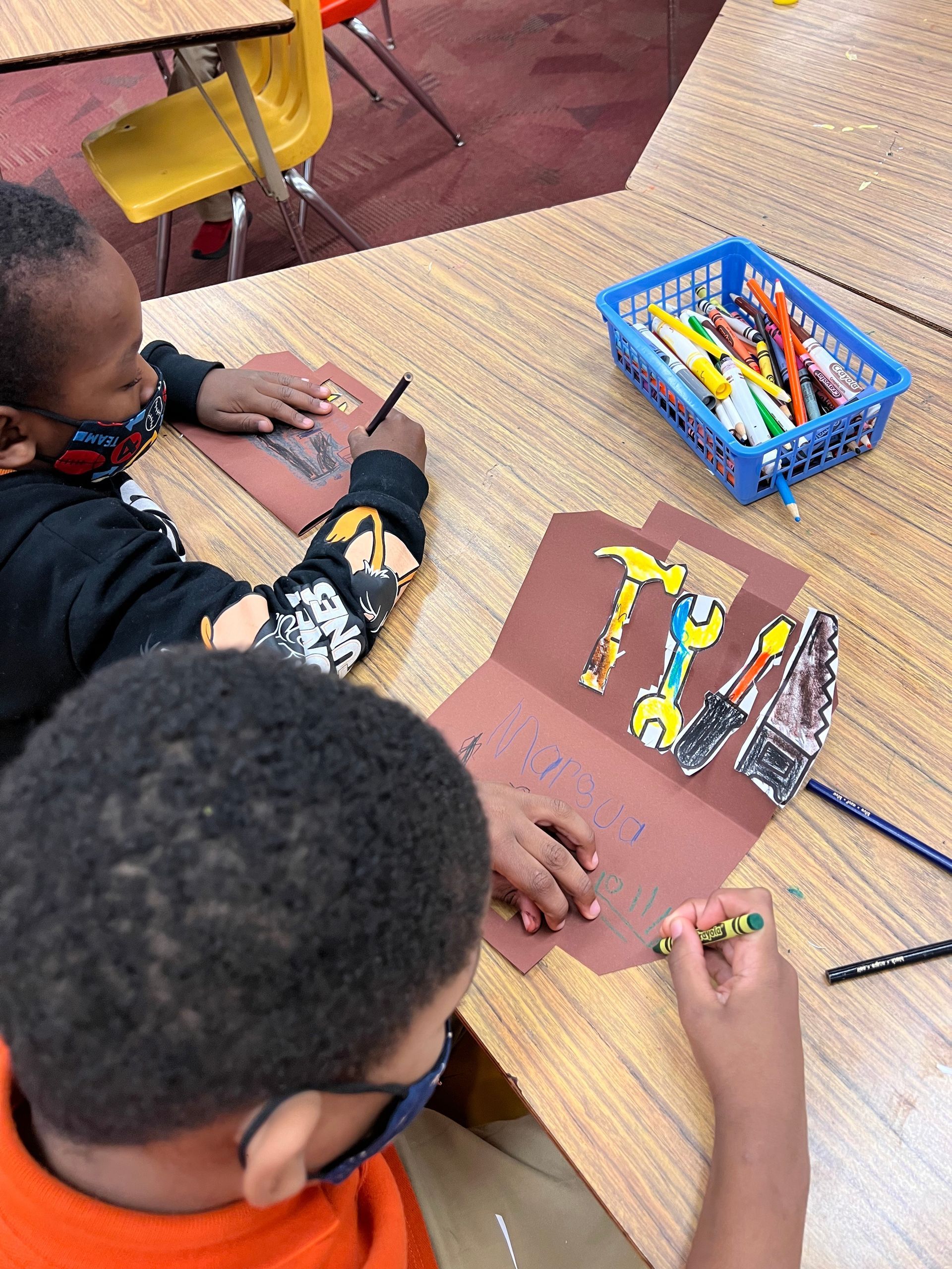 Children draw with colored pencils at a classroom table, with a blue bin of supplies nearby.