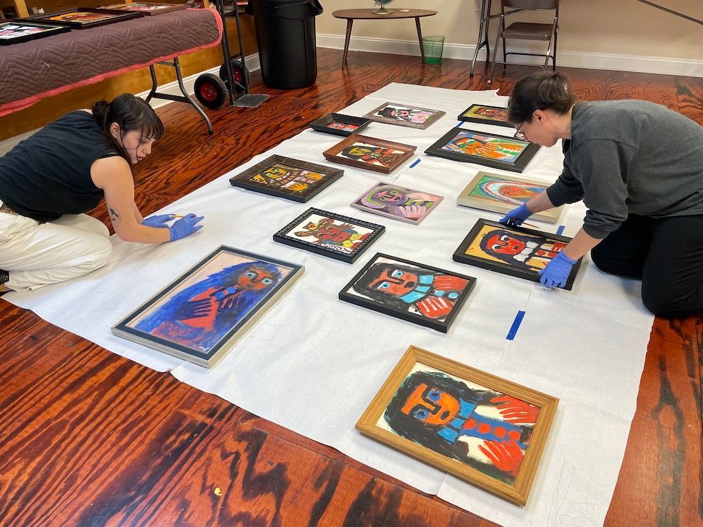 Two people arranging framed art prints on a large white sheet in a room with a wood floor.