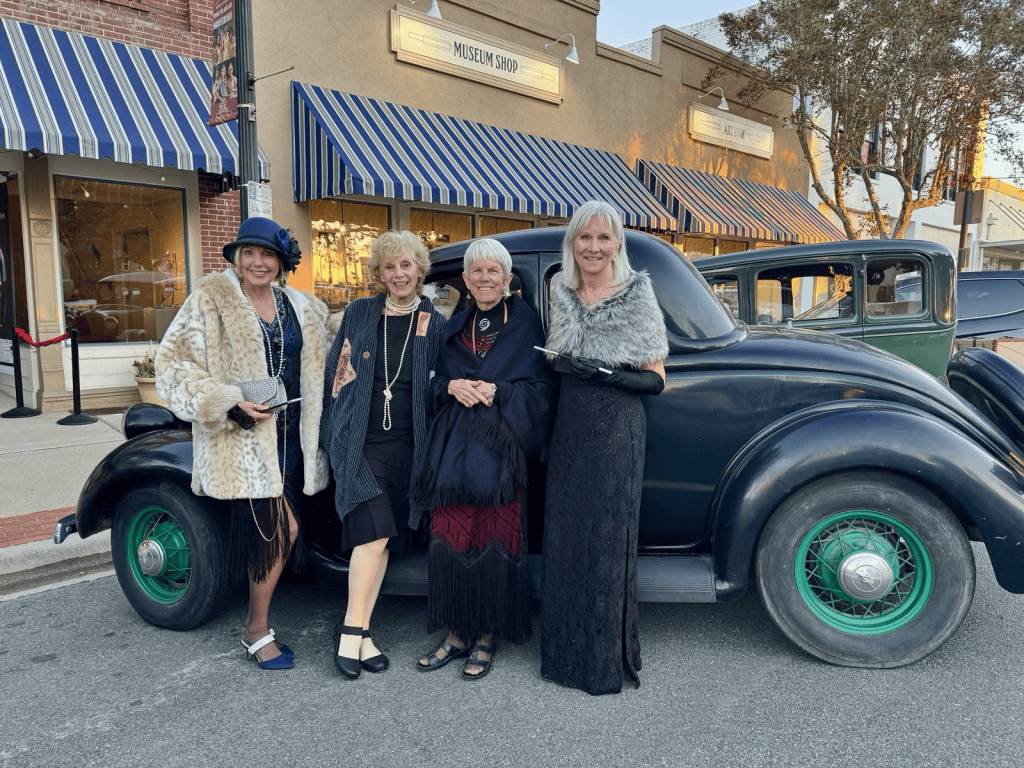 Four women standing beside a vintage black car with green wheels on a street with striped awnings