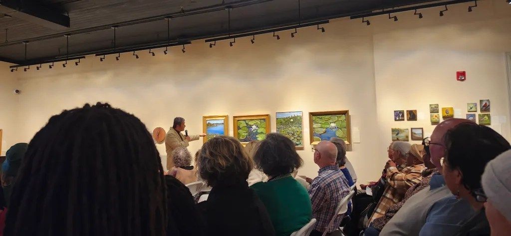 Audience seated in an art gallery, viewing framed landscape paintings on a warm-lit wall