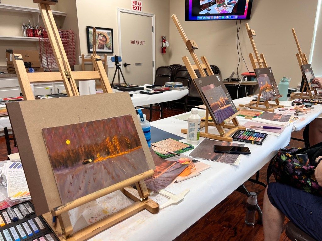 Art class table with multiple easels, paintings, and art supplies in a studio setting
