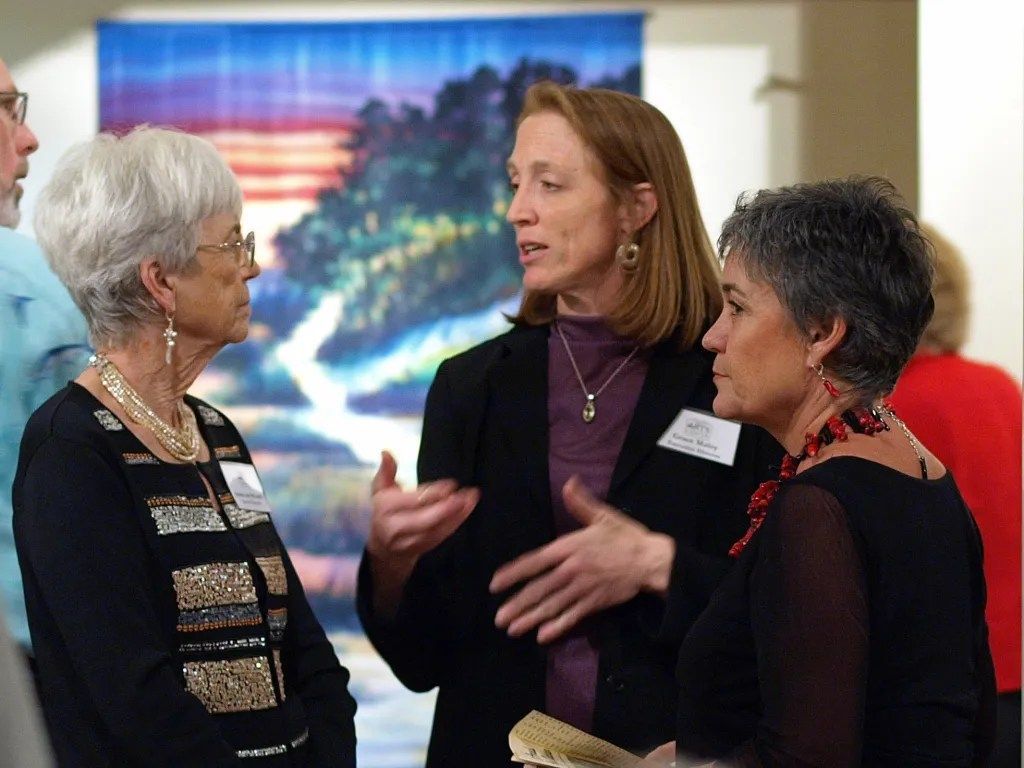 Three people talk in a bright room, with one woman gesturing while others listen.