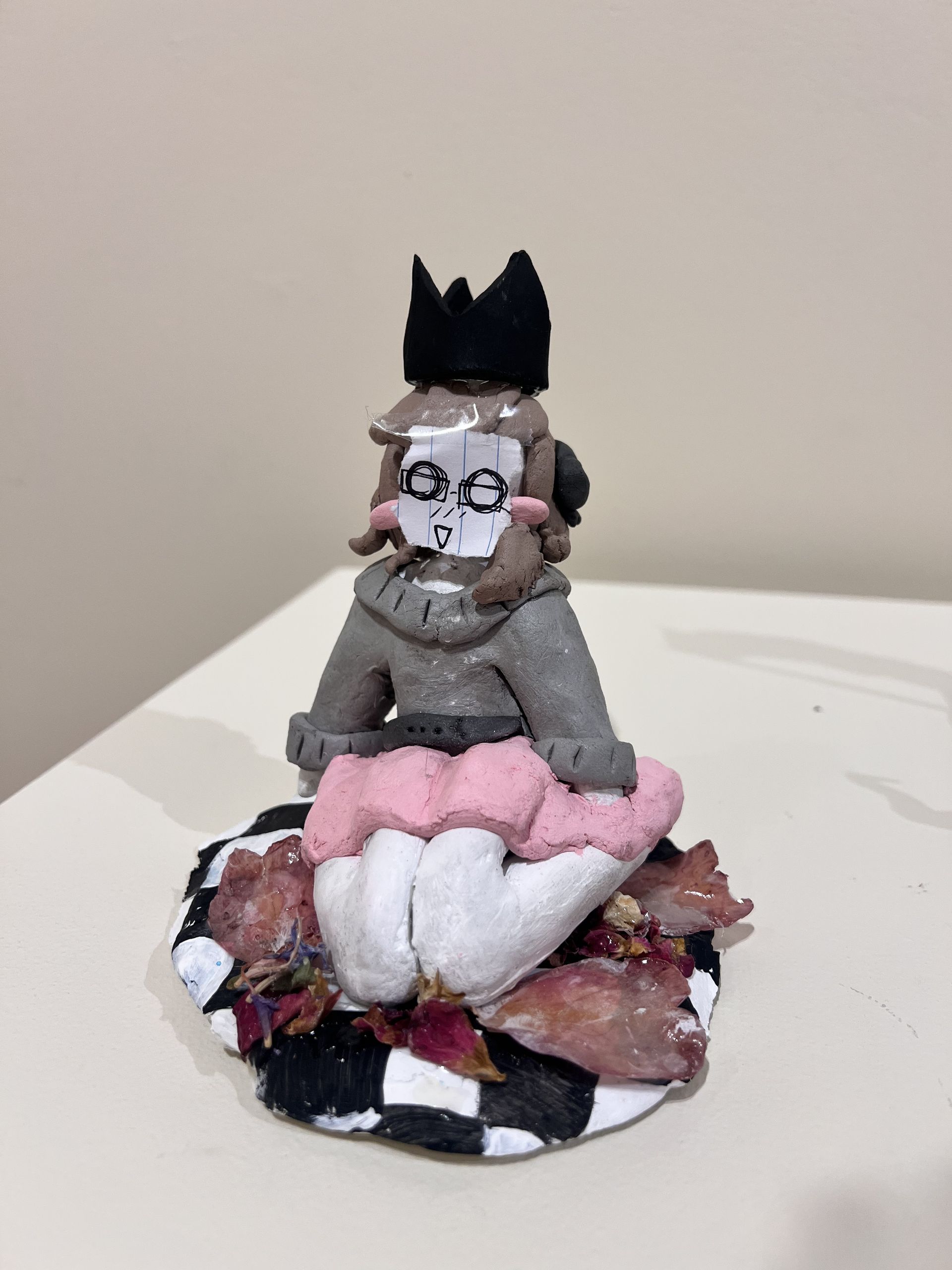 A small handmade figurine with a black crown sits on a layered pink, white, and brown flower-like base.
