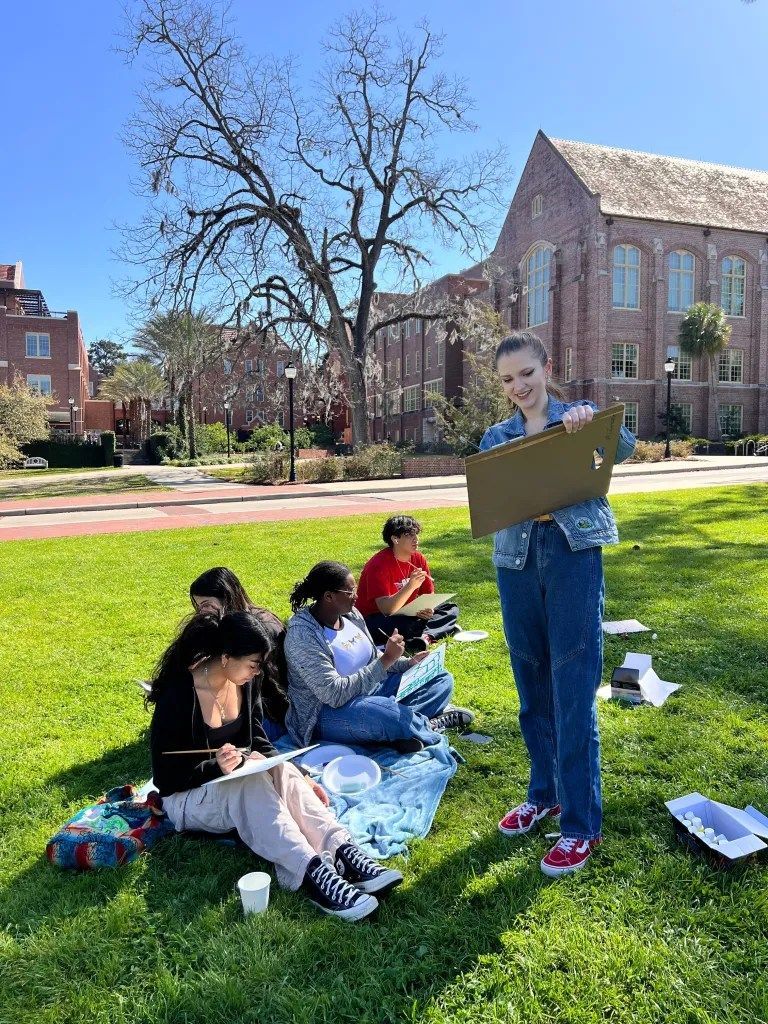 Students sitting on grass outdoors, with one person standing and holding a clipboard near a brick building.