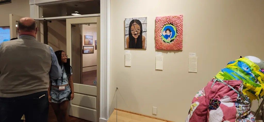 Art gallery hallway with two framed portraits on a wall and people walking past an open doorway.