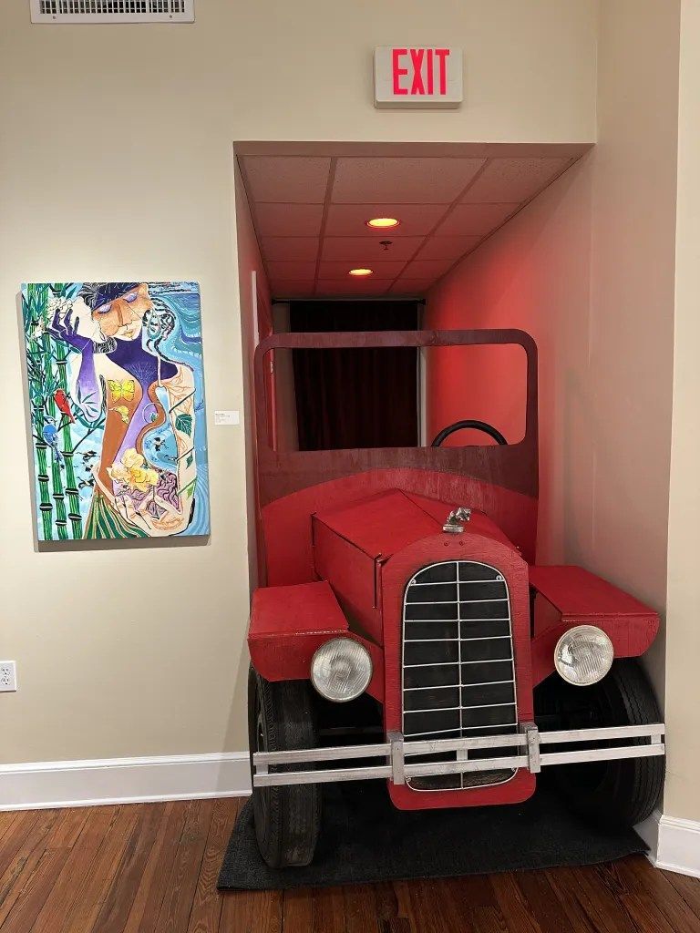 Red vintage truck displayed indoors beneath an exit sign, framed by a doorway and wall art.