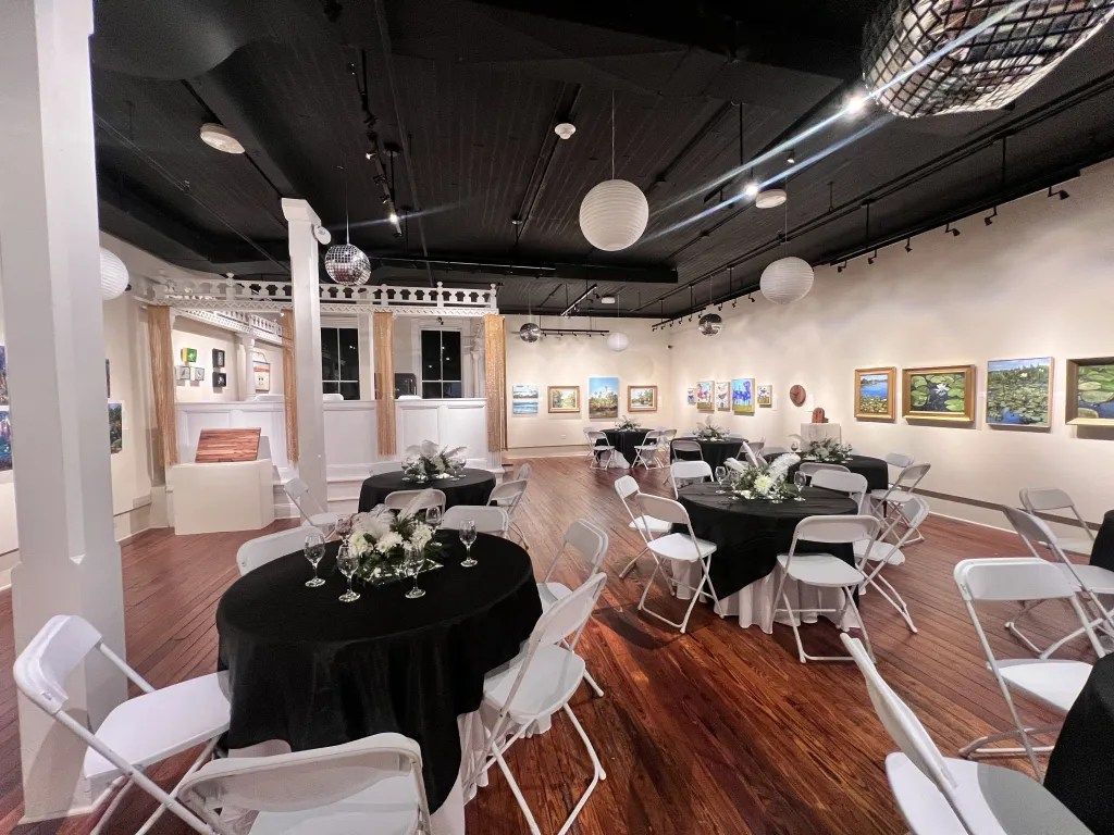 Art gallery event space with framed paintings, round tables, and white chairs under dark ceiling lights