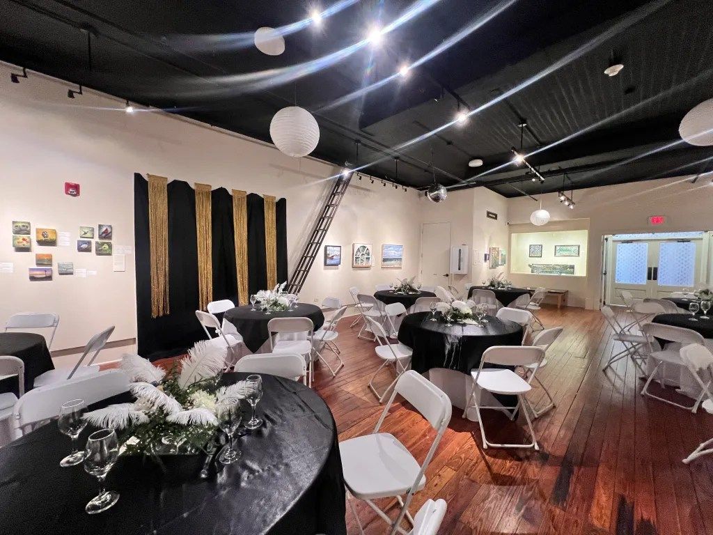 Banquet hall with round tables, white chairs, black tablecloths, and string lights on a wooden floor