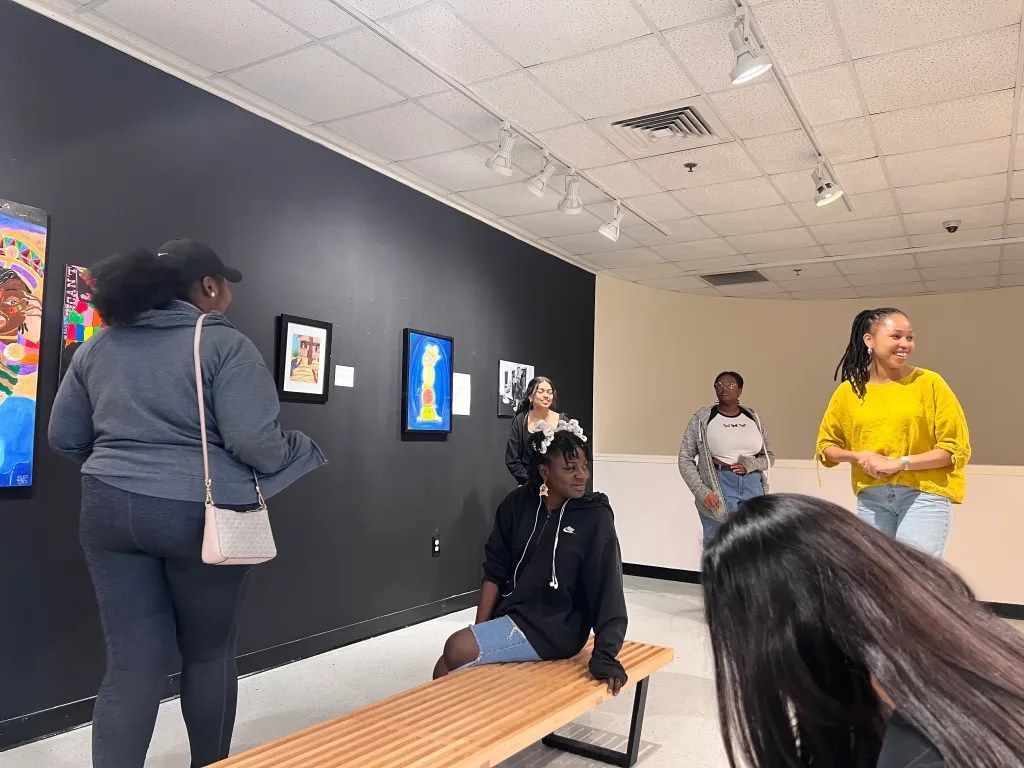 People viewing art in a gallery, with framed pieces on a dark wall and a bench in the foreground