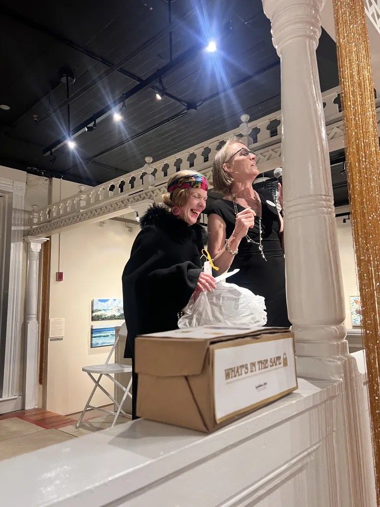 Two people standing on a raised platform in a gallery, one leaning over a cardboard box labeled “Pine…”.