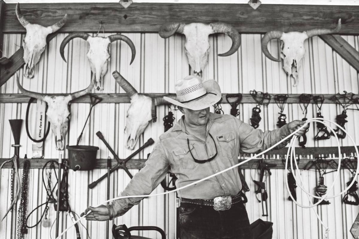 Cowboy holding a lasso in a tack room, surrounded by saddles and hanging ropes.