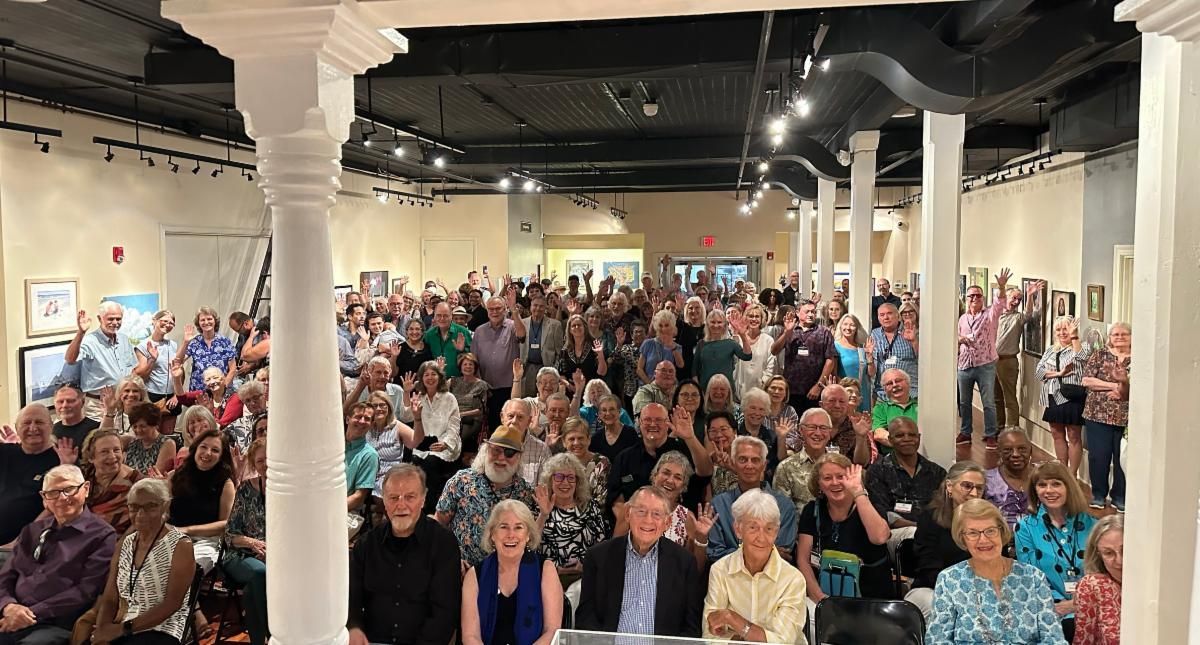 Large crowd seated in a hall, facing the camera amid white columns and industrial ceiling lights.