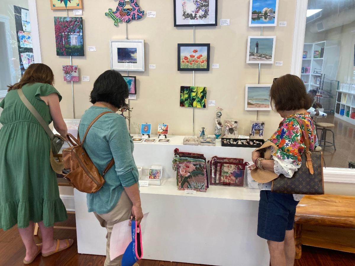 Three people browsing framed art and pottery in a bright gallery boutique