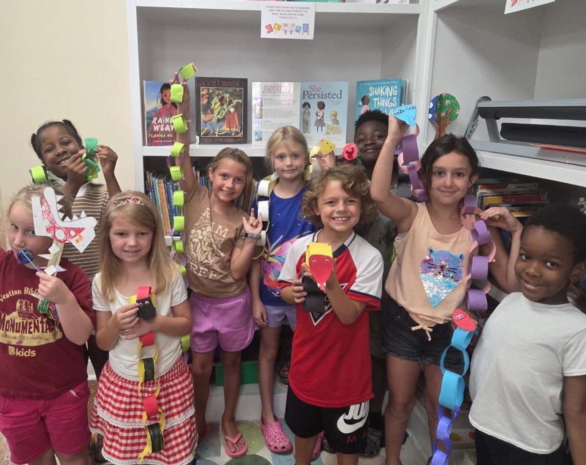Children smiling and holding colorful paper crafts in a classroom or library.