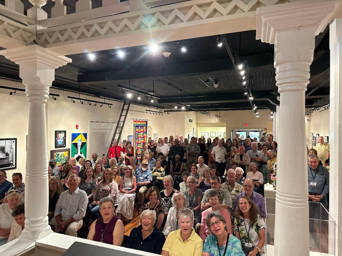 Crowded art gallery event with seated audience under bright gallery lights and white columns