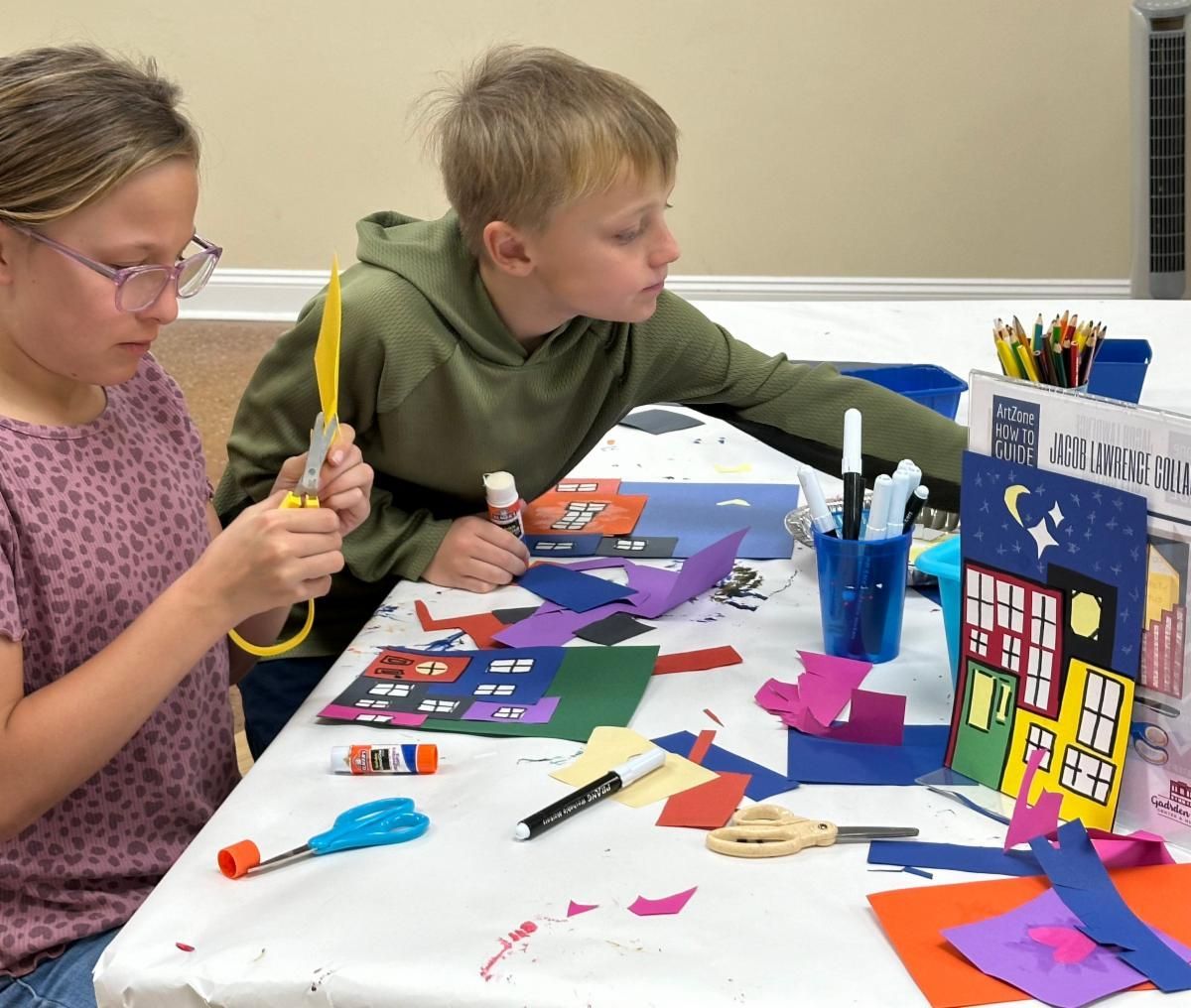 Two children craft colorful paper houses at a table with glue, markers, and scissors.