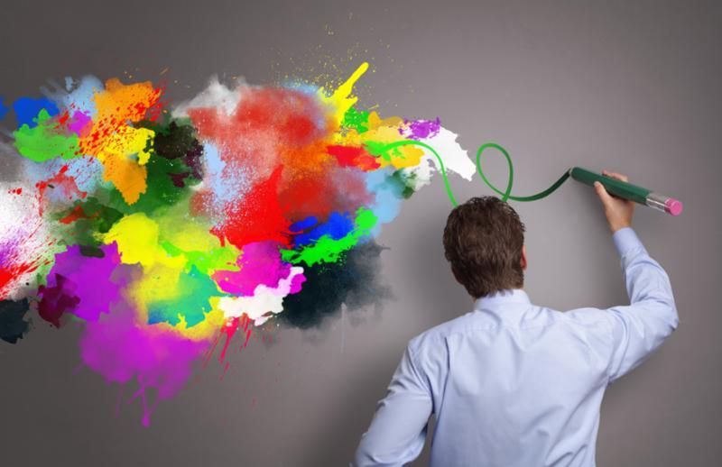 Person in a white shirt drawing a colorful abstract cloud on a gray wall with a green marker