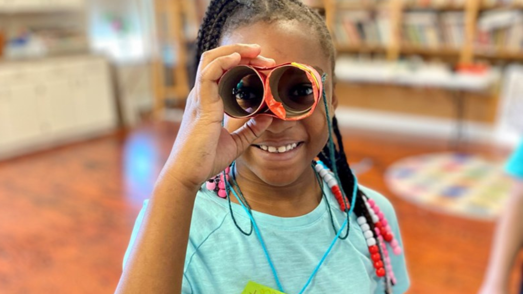 Smiling child holding orange binoculars in a library, with colorful braids and a blue shirt