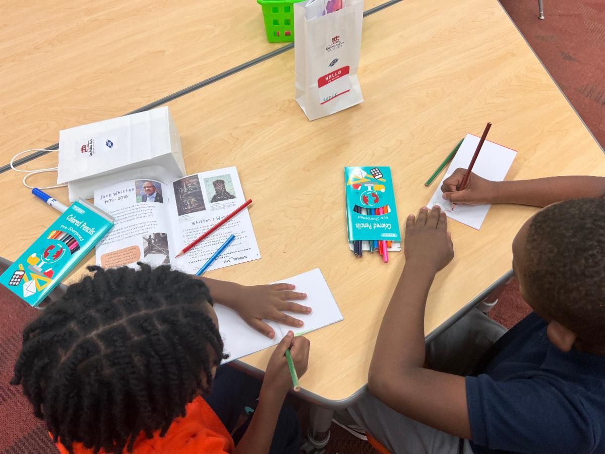 Children writing at a classroom table with books, pencils, and worksheets spread out.
