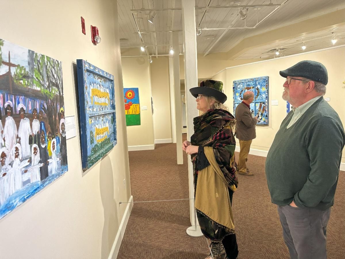 People viewing framed artwork in a gallery hallway, with white walls and a carpeted floor.