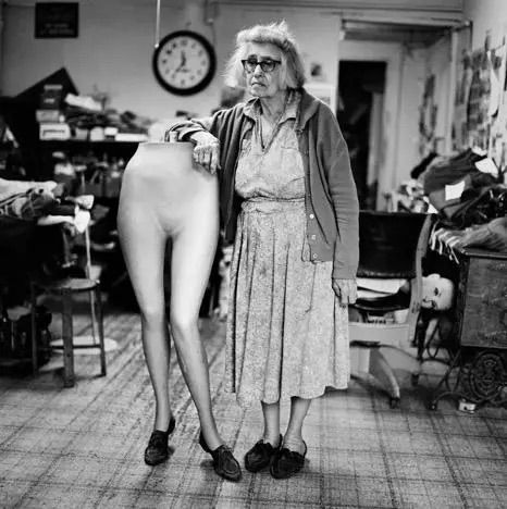 Woman in a workshop standing beside a mannequin of legs, with sewing materials and tools around her.