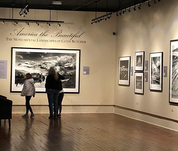 Art gallery with two visitors viewing black-and-white landscape photographs on beige walls.