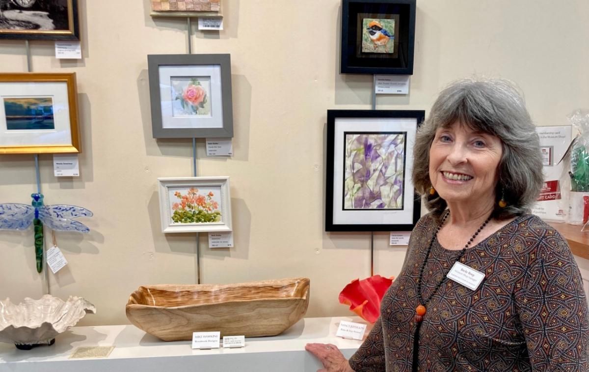 Woman standing beside framed artwork and a display table at an art exhibit