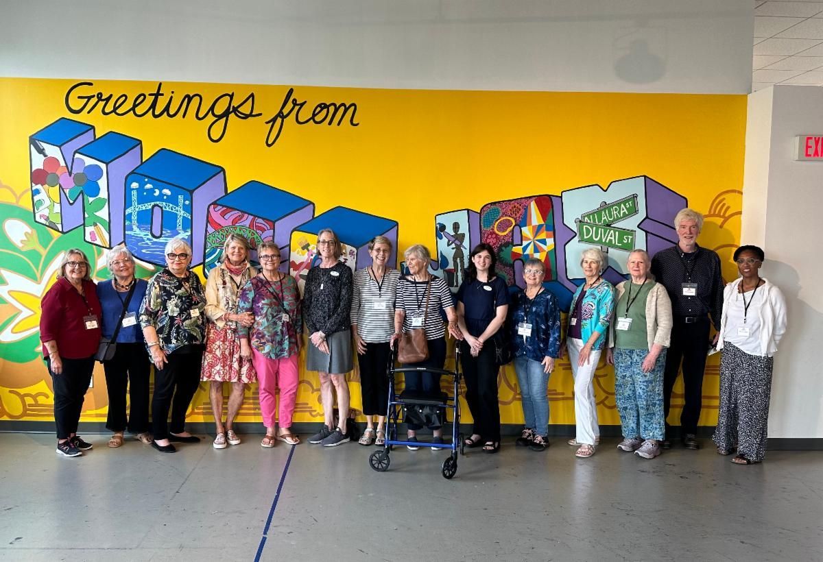 Group of people posing in front of a colorful mural with “Greetings from” text on a yellow wall.