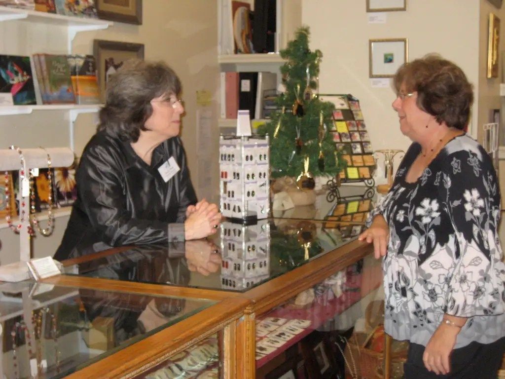 Two women converse across a glass counter beside a small Christmas tree in a shop.