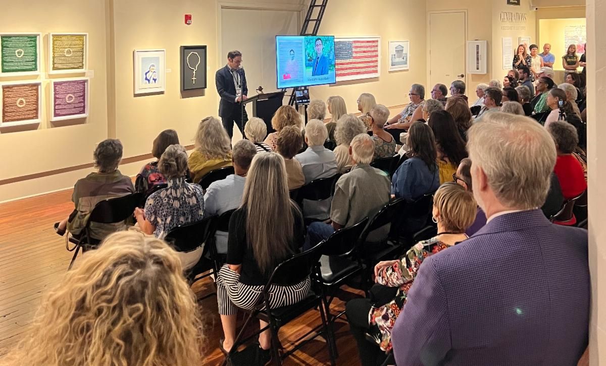 Audience seated in a gallery listening to a speaker beside a blue presentation screen