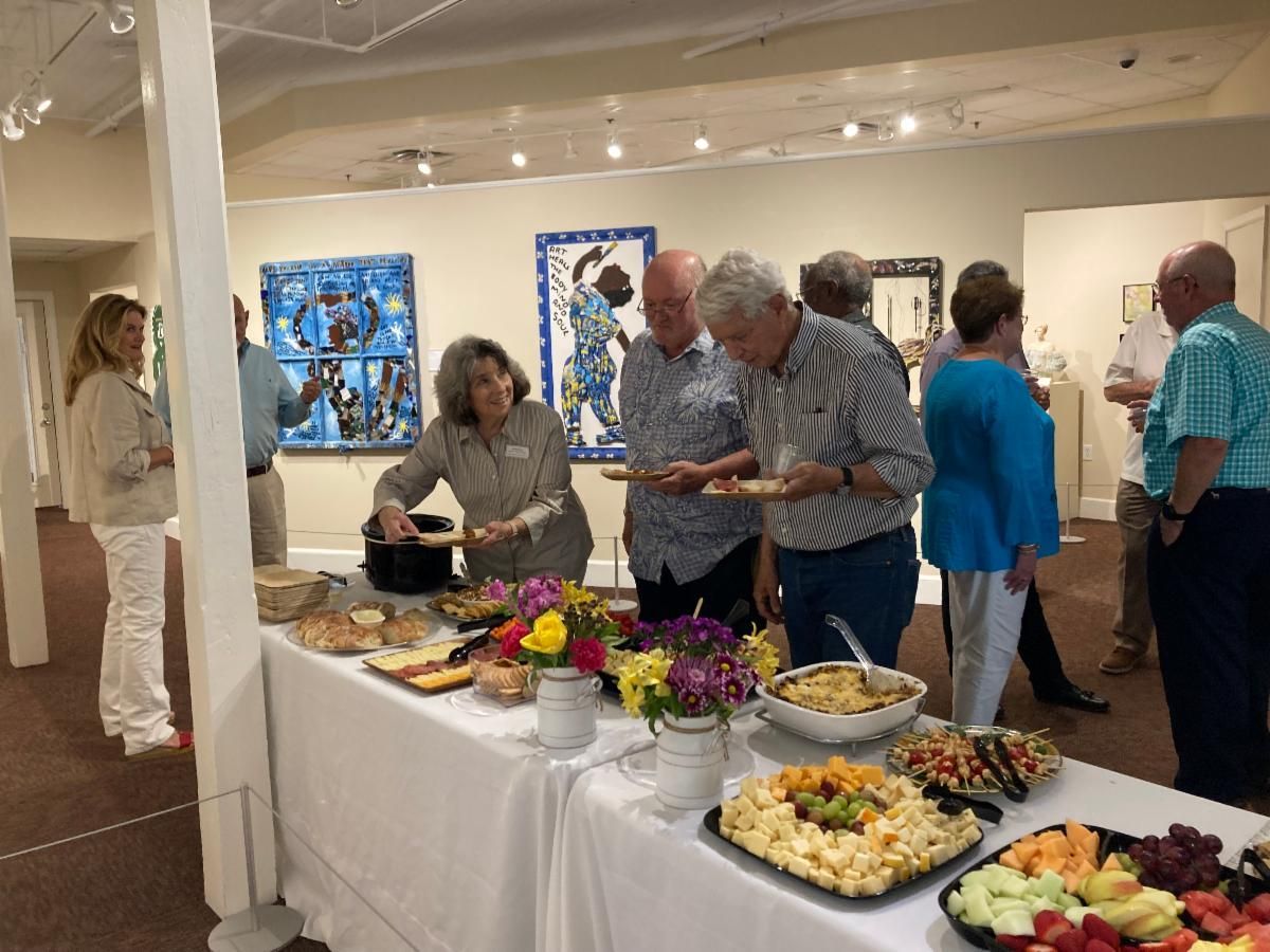 Guests chat at an art gallery buffet with food trays and flower arrangements on a white table.