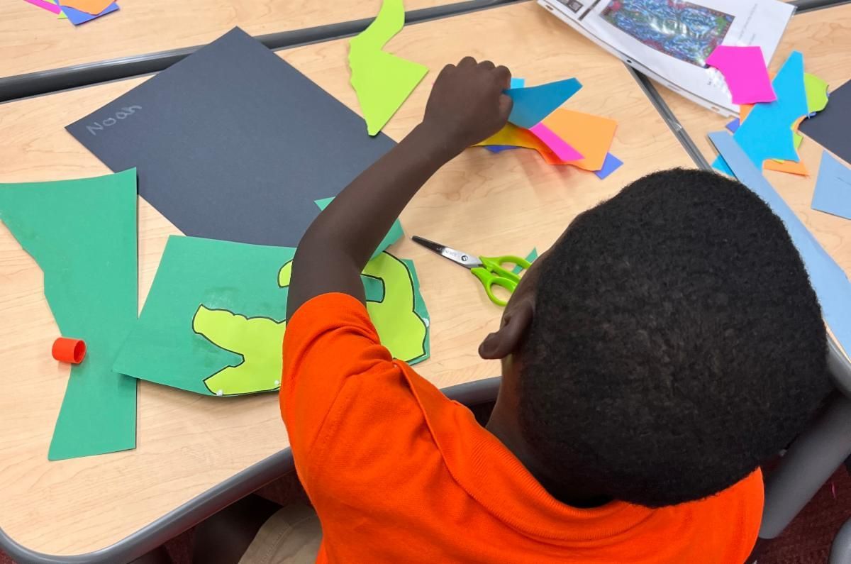 Child in an orange shirt arranging colorful paper shapes on a table for a craft activity