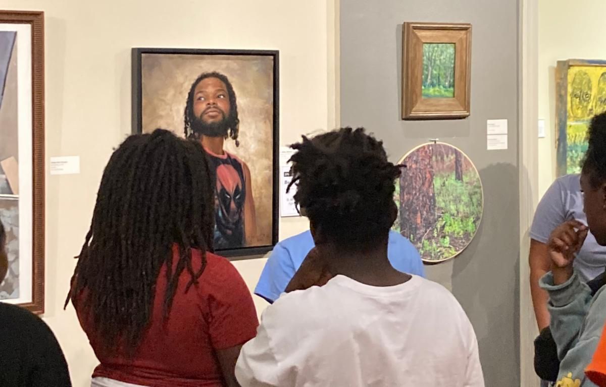 Museum visitors view framed artworks in a gallery, including a portrait and colorful abstract pieces.