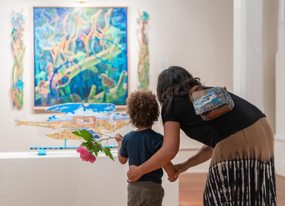 Adult and child viewing a colorful painting in a gallery, with the adult leaning toward the child.