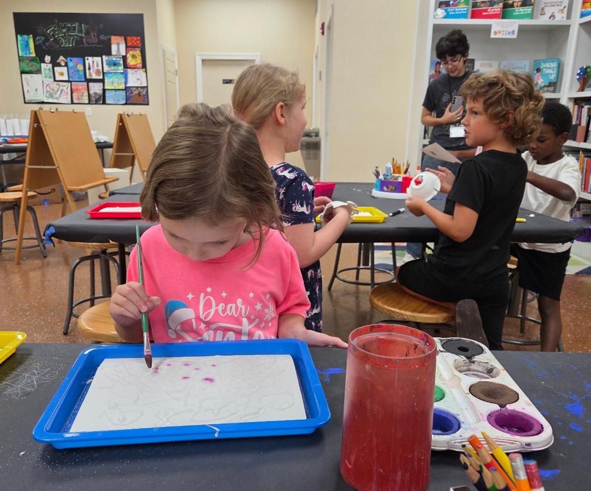 Children doing arts and crafts at a classroom table with paint, brushes, and colorful supplies.