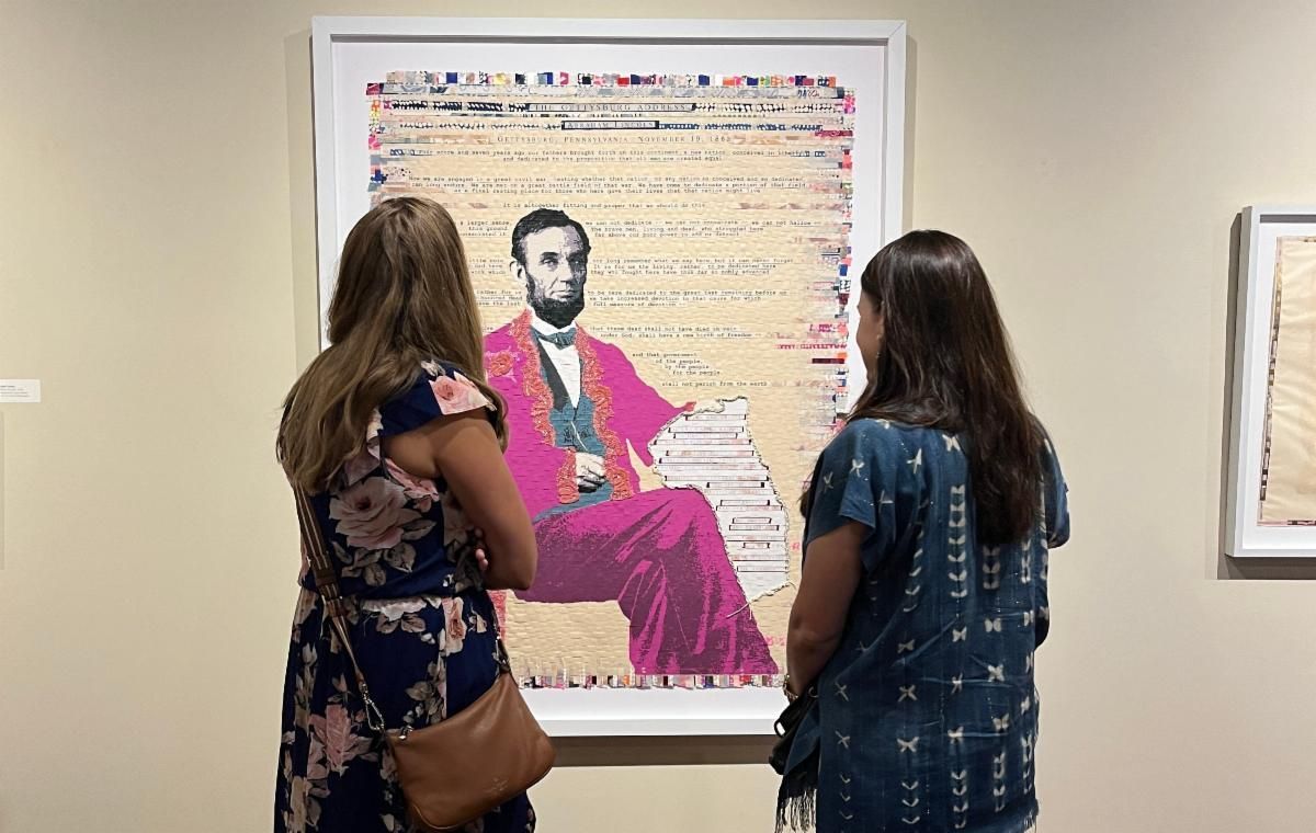 Two people view a framed portrait of Abraham Lincoln with a bright pink suit in a gallery.