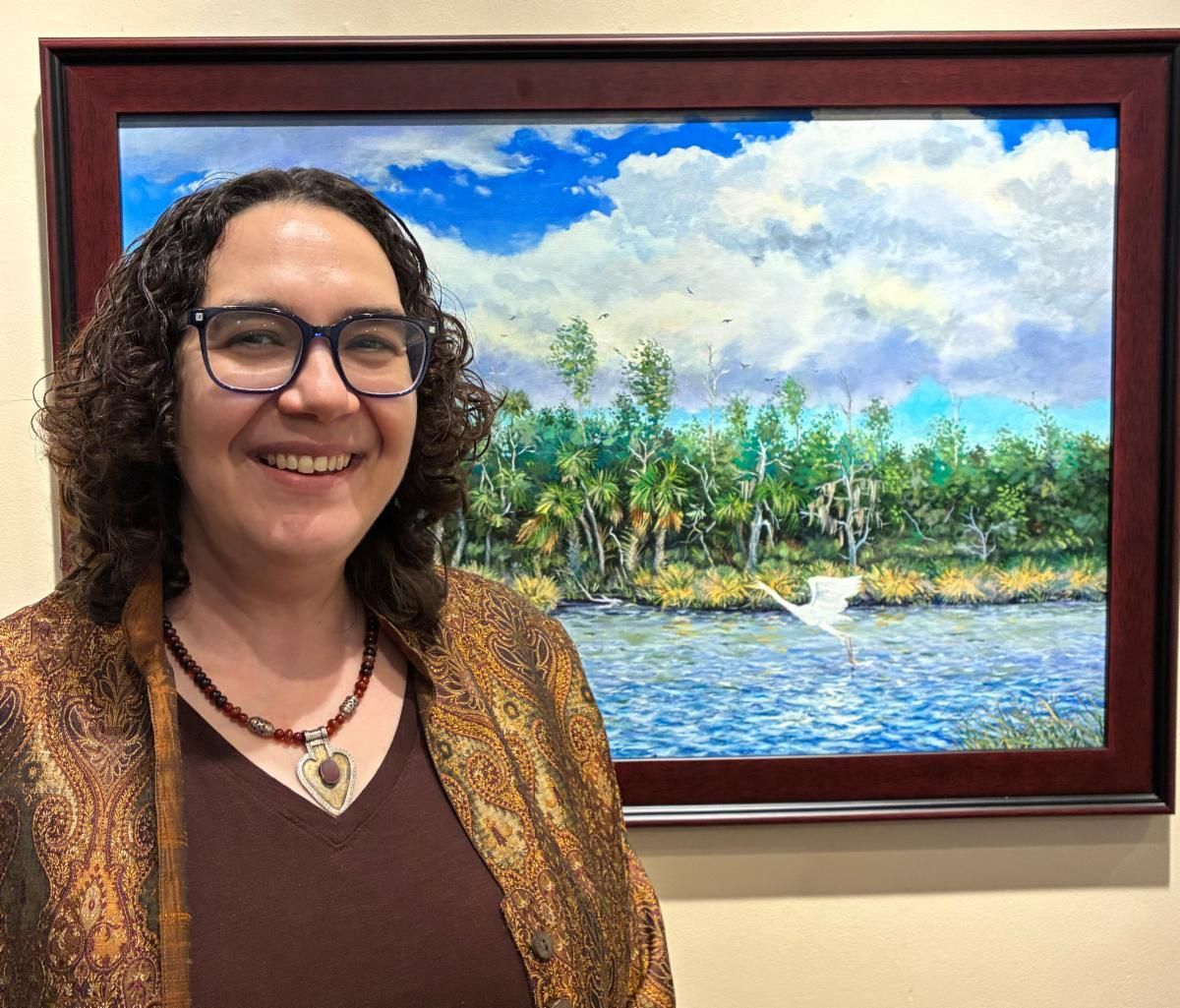 Smiling person stands beside a framed tropical landscape painting in a gallery.
