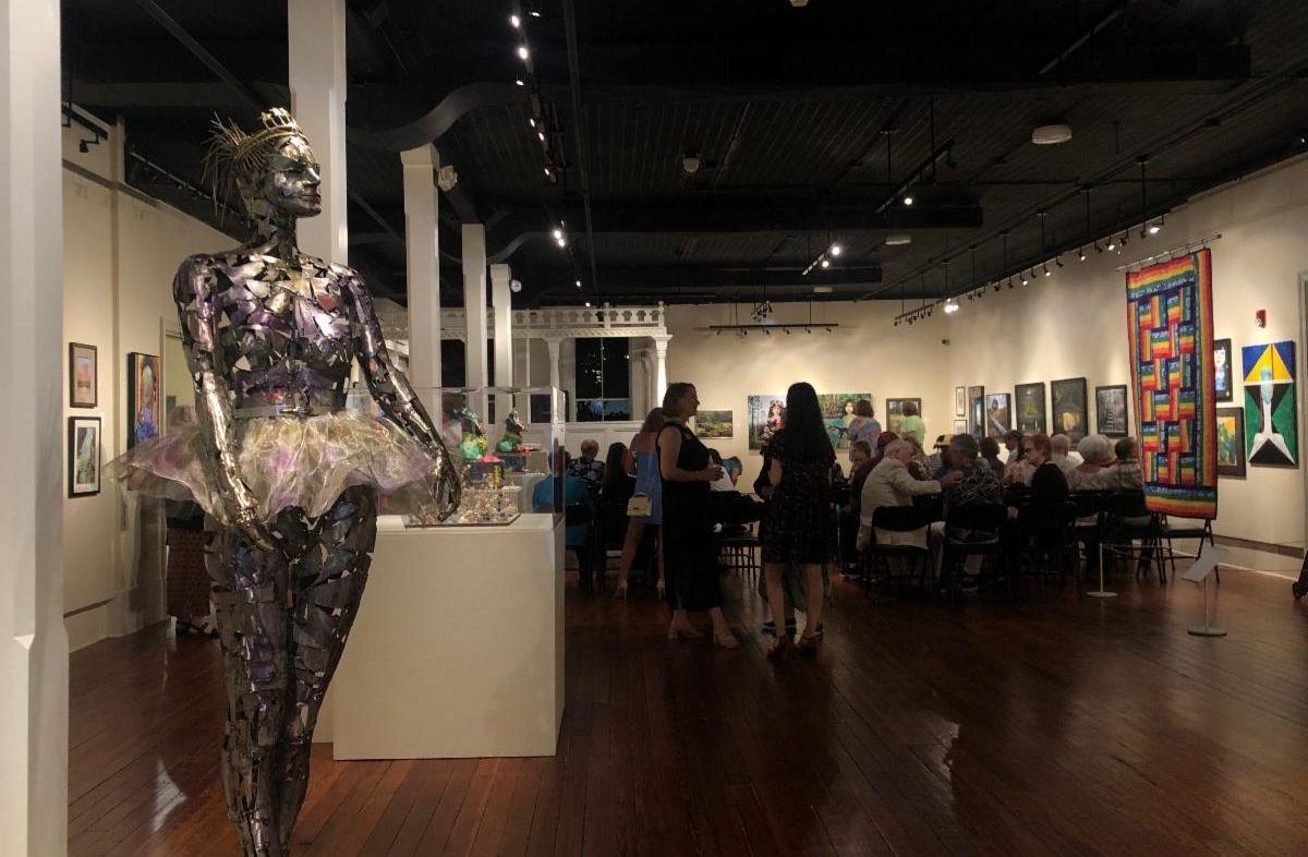 Art gallery interior with a silver sculpture in the foreground and people browsing exhibits in the background