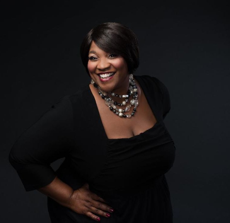 Smiling woman in black dress with statement necklace, posing against a dark background