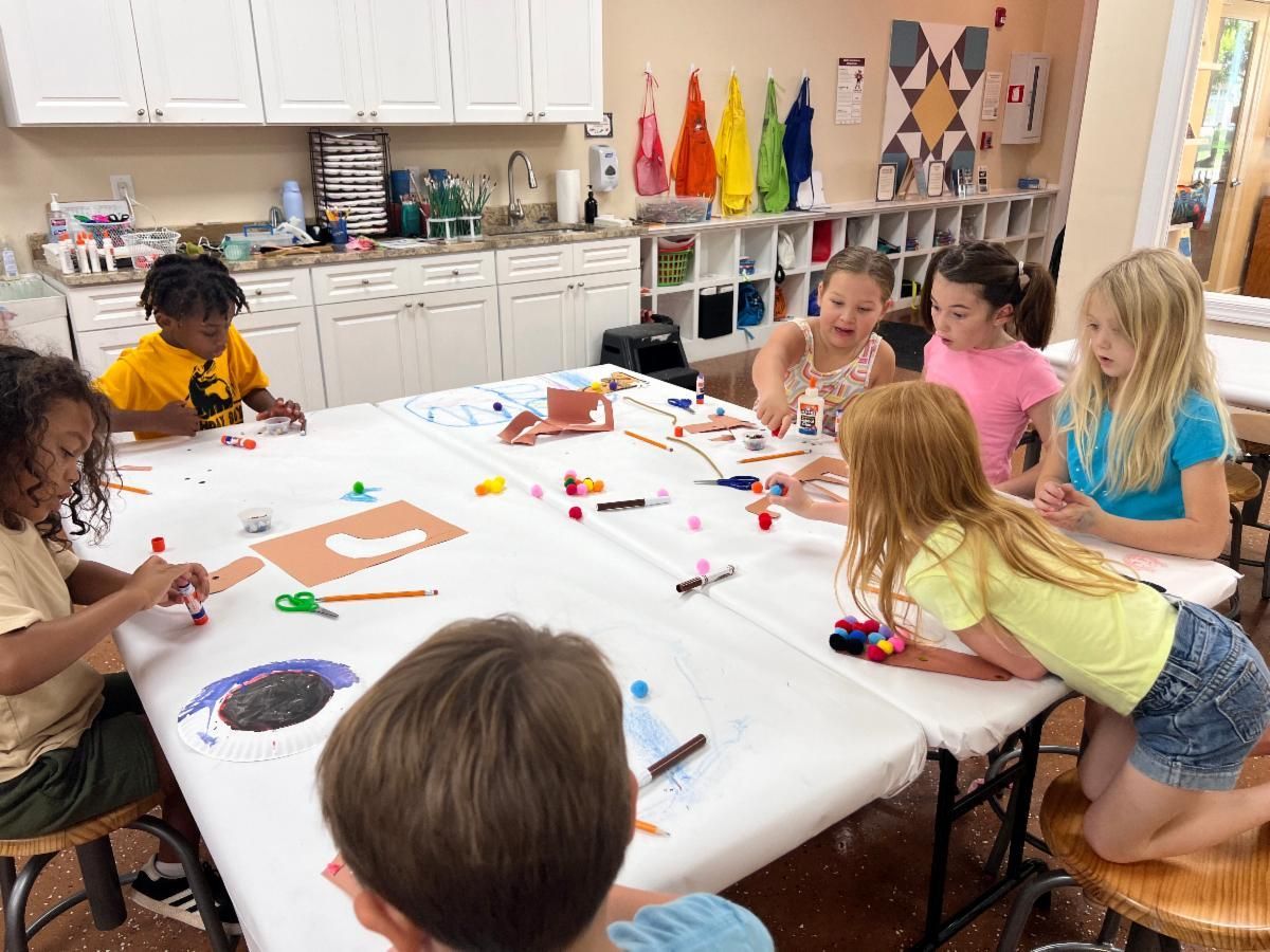 Children painting at a classroom table with colorful art supplies and paper crafts