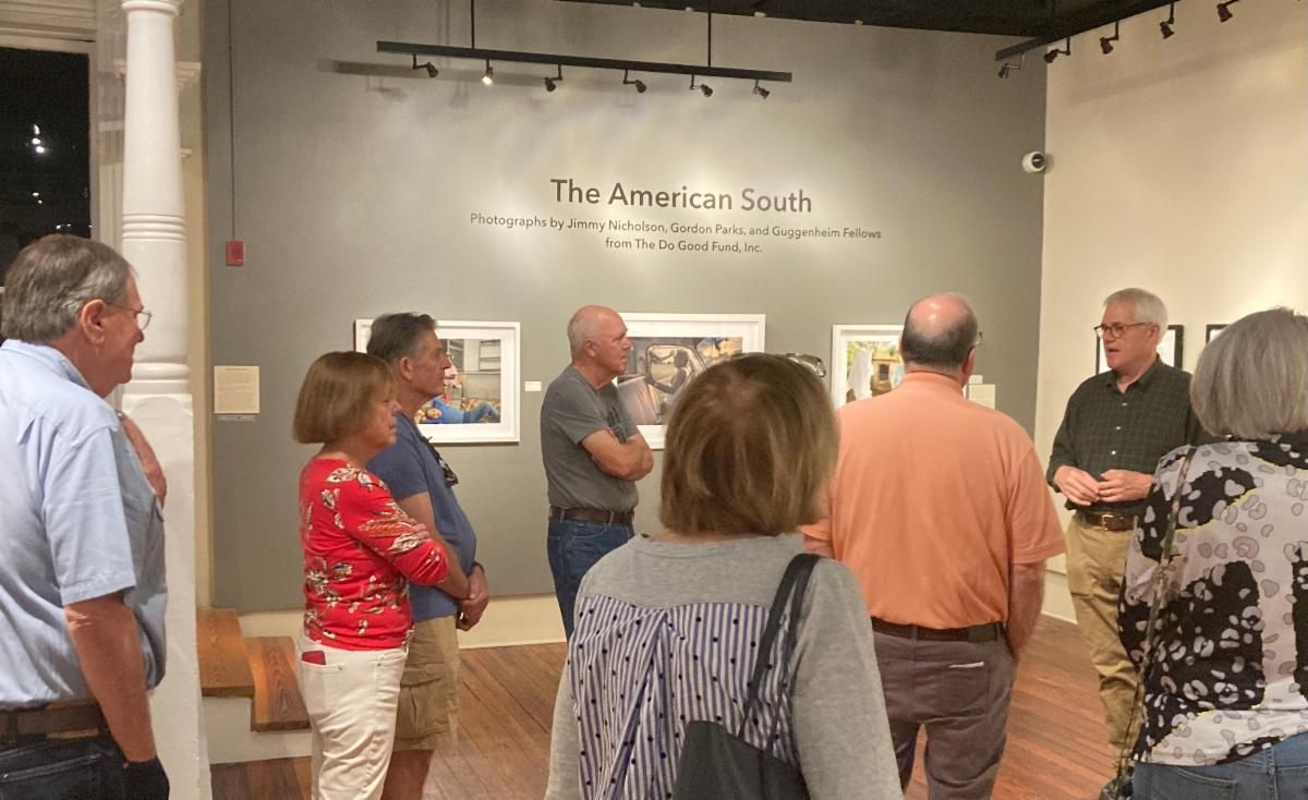 People gathered in a gallery, listening to a speaker beside wall text and an artwork display.