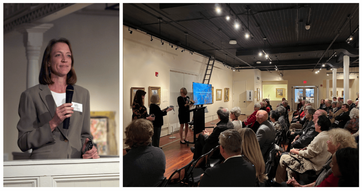 Woman speaking at a podium beside a seated audience in an art gallery, with a screen and microphone visible.