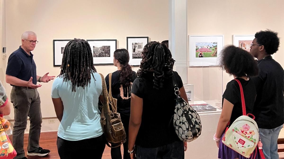 Visitors listening to a guide in an art gallery, viewing framed photos on a white wall.