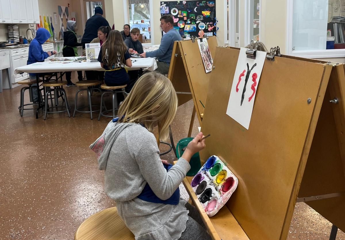 Child painting at an easel in a classroom, with art supplies and other students in the background